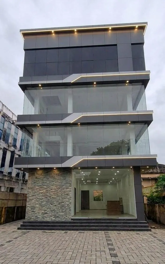 Four-storey commercial building with a glass facade and a lit cornice, on a paved forecourt