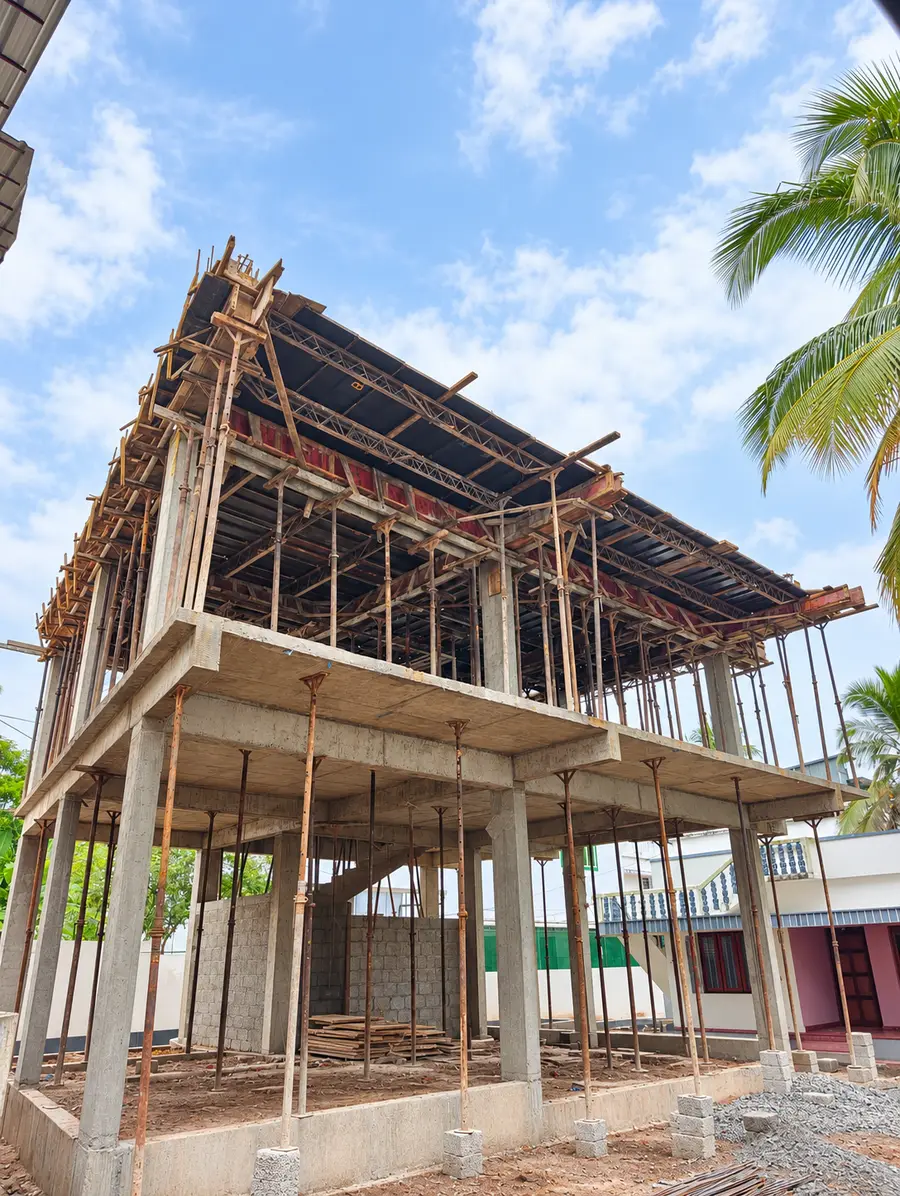 Two-storey concrete frame under construction with timber and steel props holding up the roof slab, a palm tree and a white house beside it
